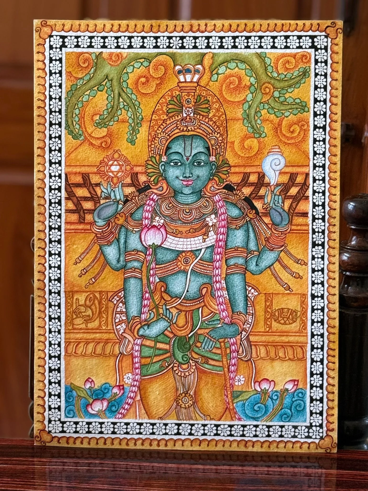 A green-skinned, four-armed deity in ornate gold jewelry and a tall crown, holding a chakra and a conch, standing above a pond of pink lotus flowers with a long pink garland draped to the feet.