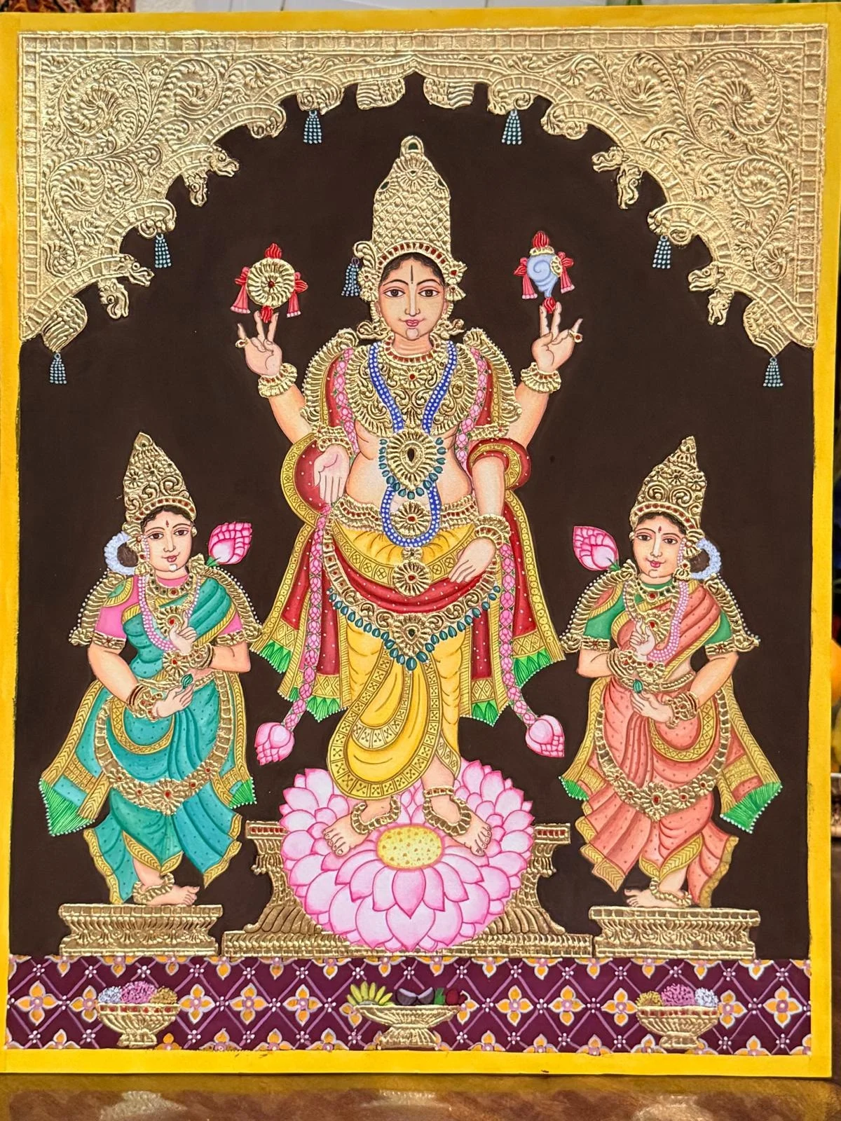 Goddess Lakshmi in raised gold-relief work, standing on a pink lotus beneath a gold-embossed arch, flanked by two female attendants in teal and pink sarees against a dark maroon background.