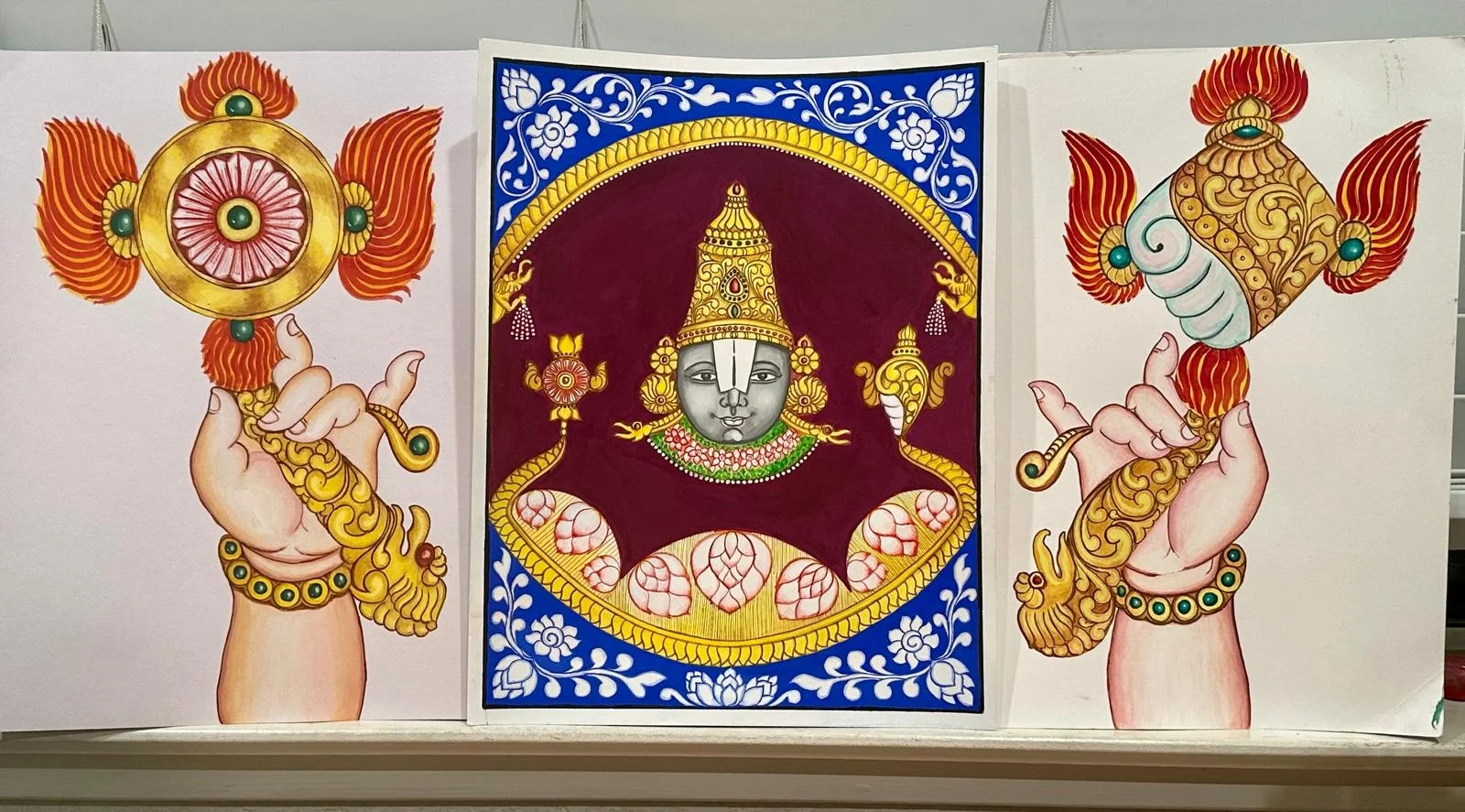 Three folk paintings displayed together: an ornamented golden discus (chakra) held in a hand on the left, a devotional close-up portrait of Balaji's face in gold and blue at center, and a golden conch (shankha) held in a hand on the right.